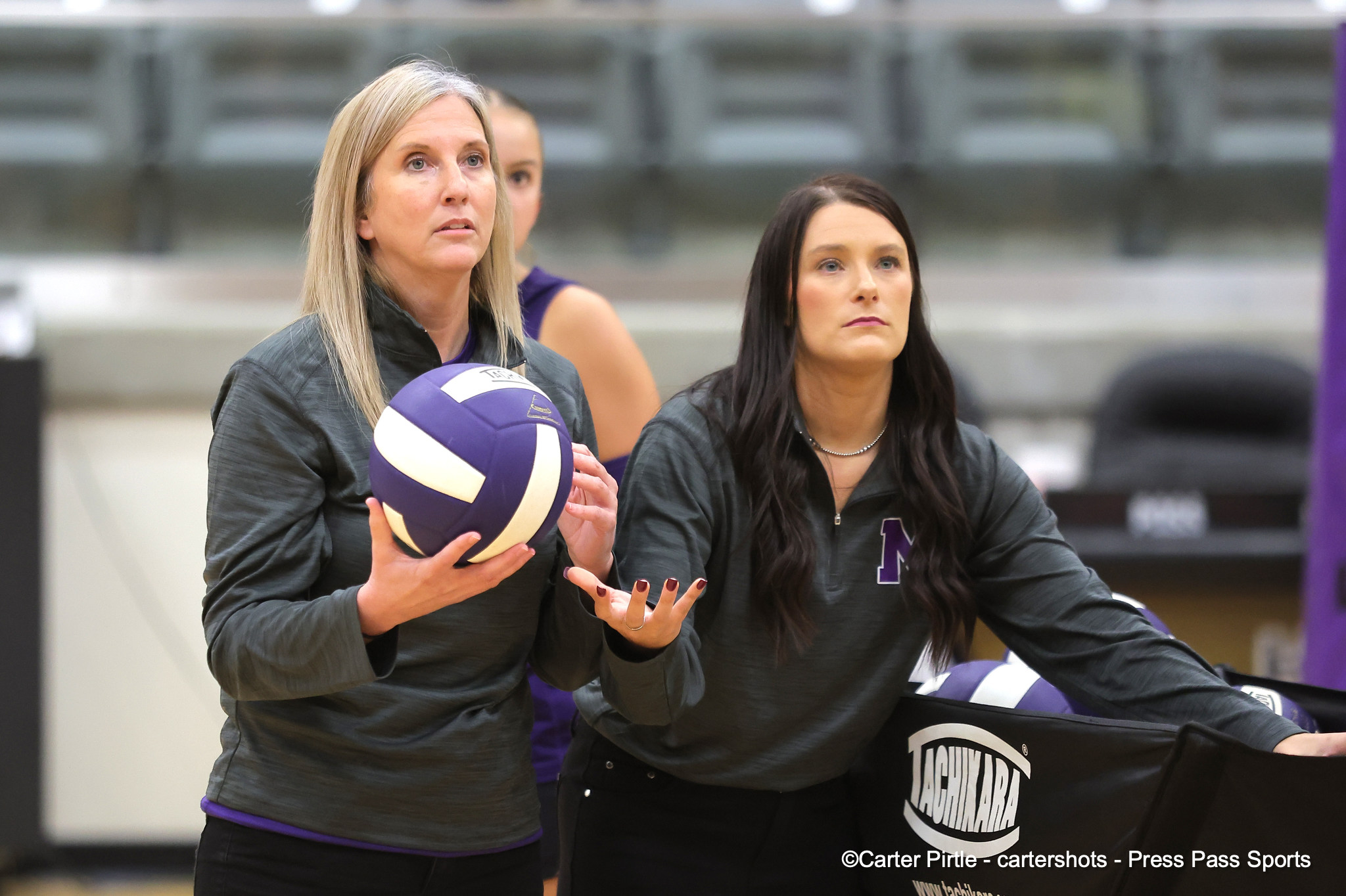 Two female volleyball players in gray tops practice in a gym; the blonde player holds a purple and white volleyball while the other watches reached hands ready for play.