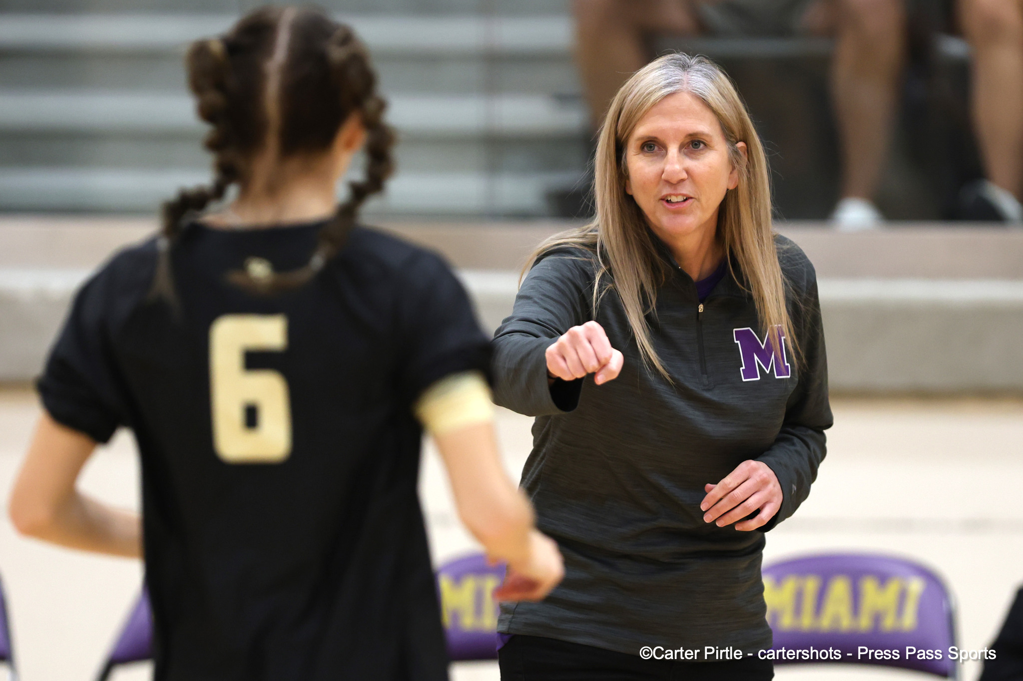 Two female athletes fist-bumping in a gym during practice; blonde woman in a gray top with a purple'M' on her chest reaches out as she greets a teammate in black behind her.