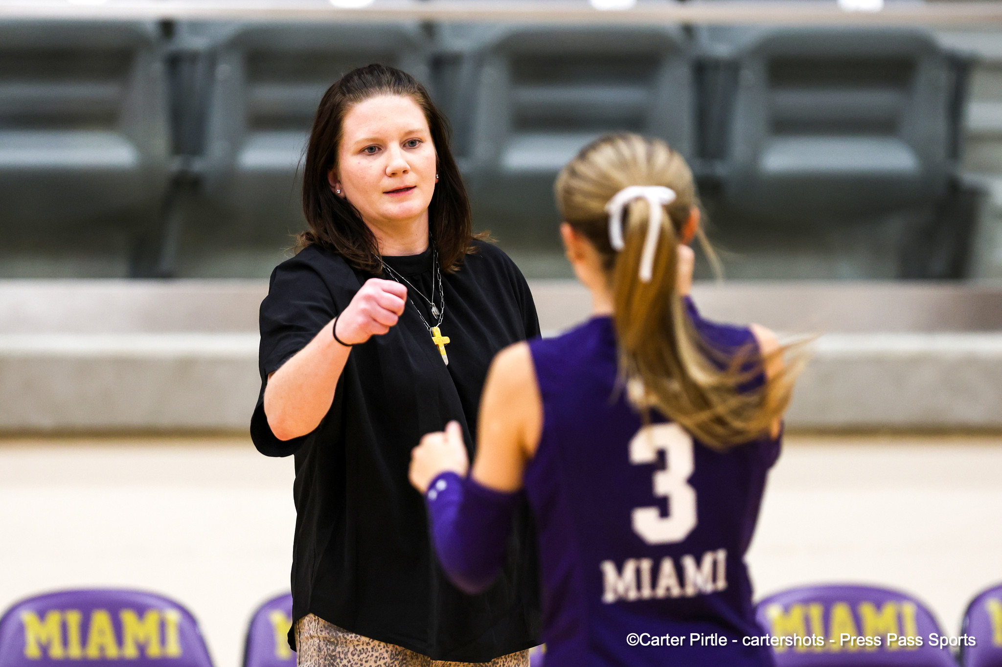 Coach or adult woman in black shirt giving a high-five to a young athlete in a purple Miami jersey in a gymnasium; bleachers in the background.