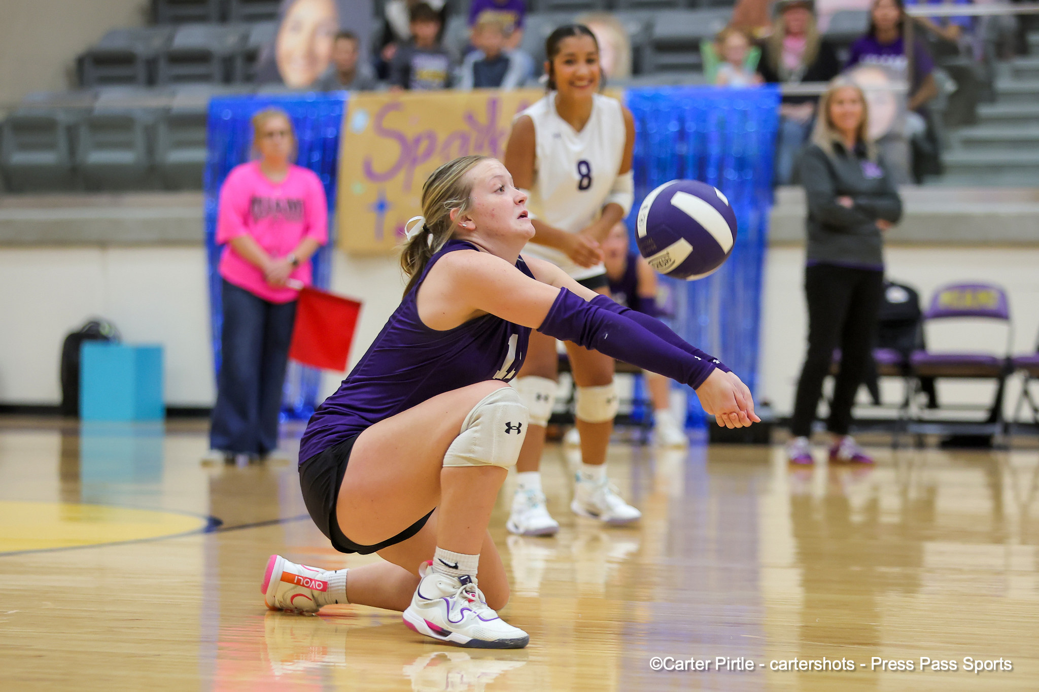 Female volleyball player in purple dives for the ball on a polished gym floor as teammates and spectators watch from the stands behind a blue backdrop.