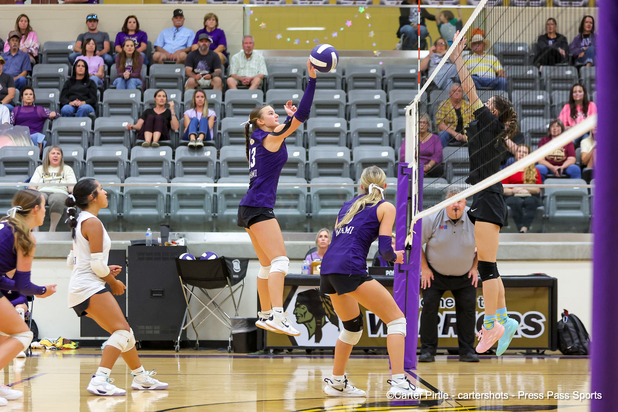 Female volleyball players in purple uniforms jump at the net to block/return the ball as fans watch from the crowded bleachers in a gym.