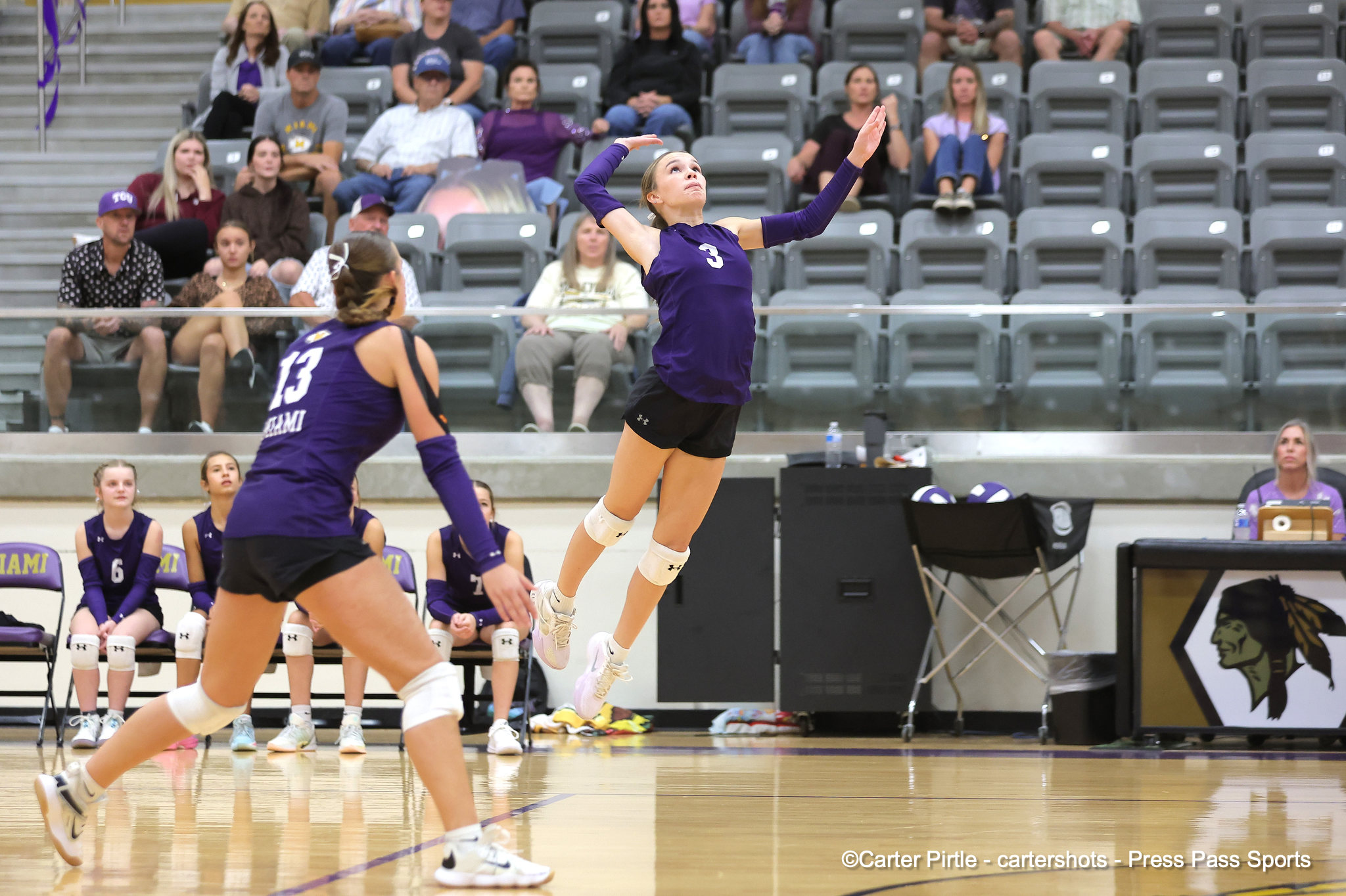 Female volleyball player in a purple jersey (#3) leaps mid-air for a spike during a match, with teammates and spectators in the stands behind her.
