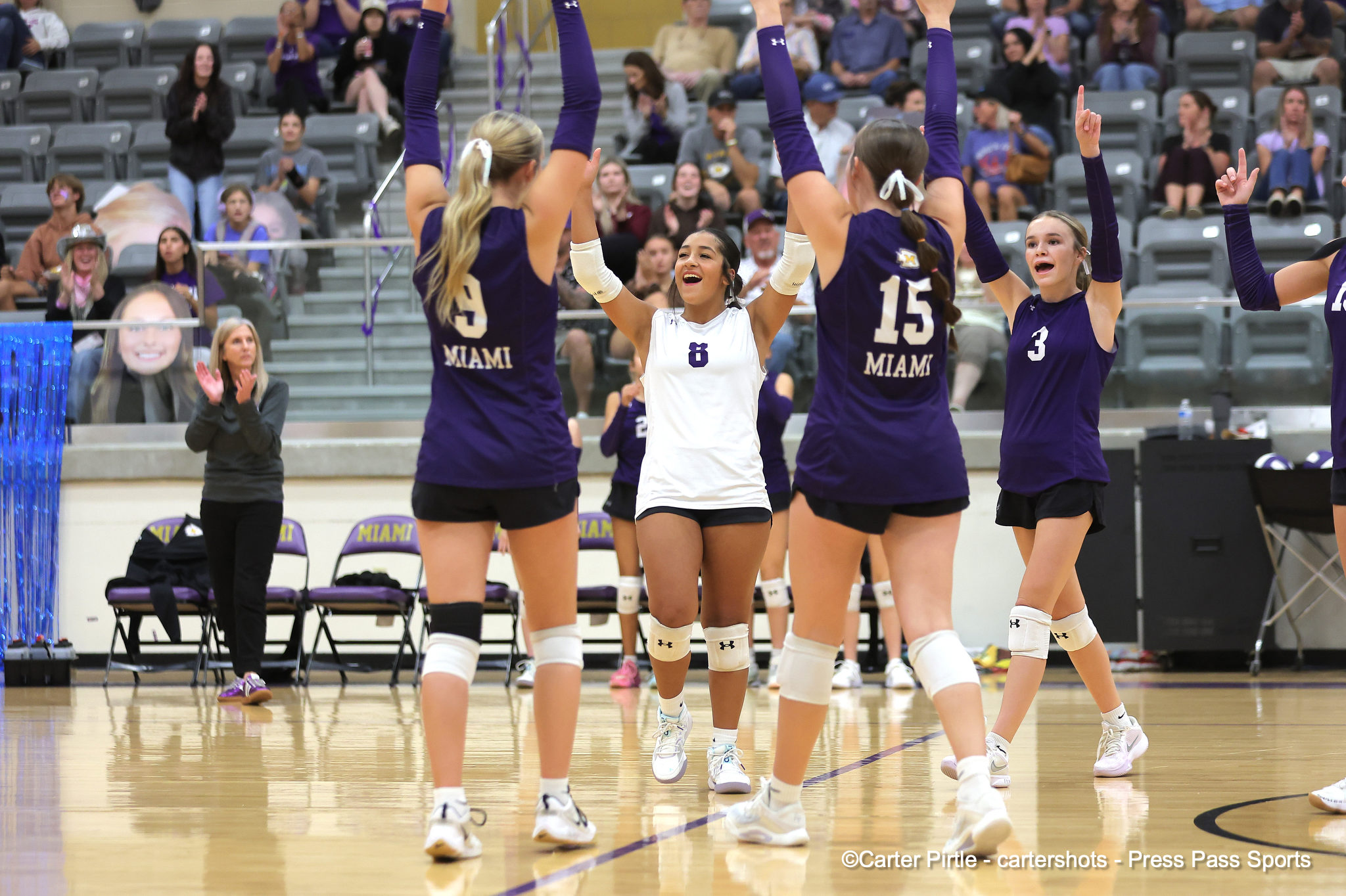 Female volleyball players celebrate with raised arms on the court; purple Miami uniforms surround a smiling white-jersey player (#6).