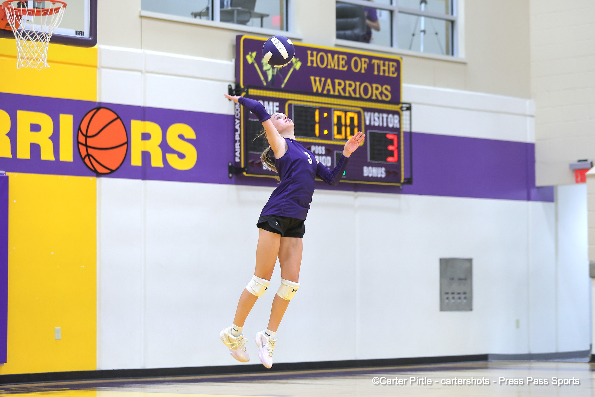 Volleyball player in a purple uniform leaps to spike the ball in an indoor gym; scoreboard in the background says'Home of the Warriors'.