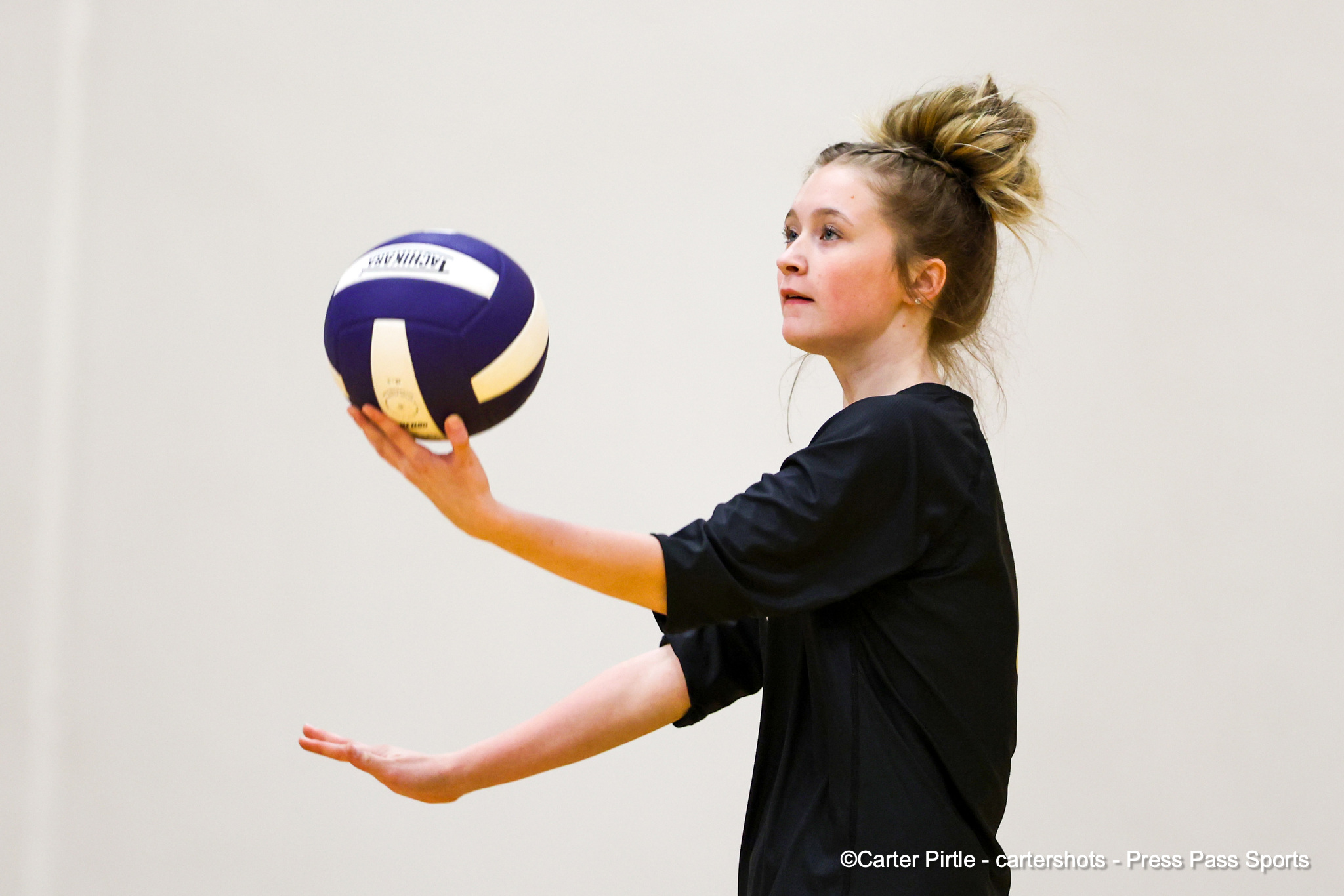 Female volleyball player in a black shirt prepares to serve with a blue and white volleyball in a gym feel indoors.