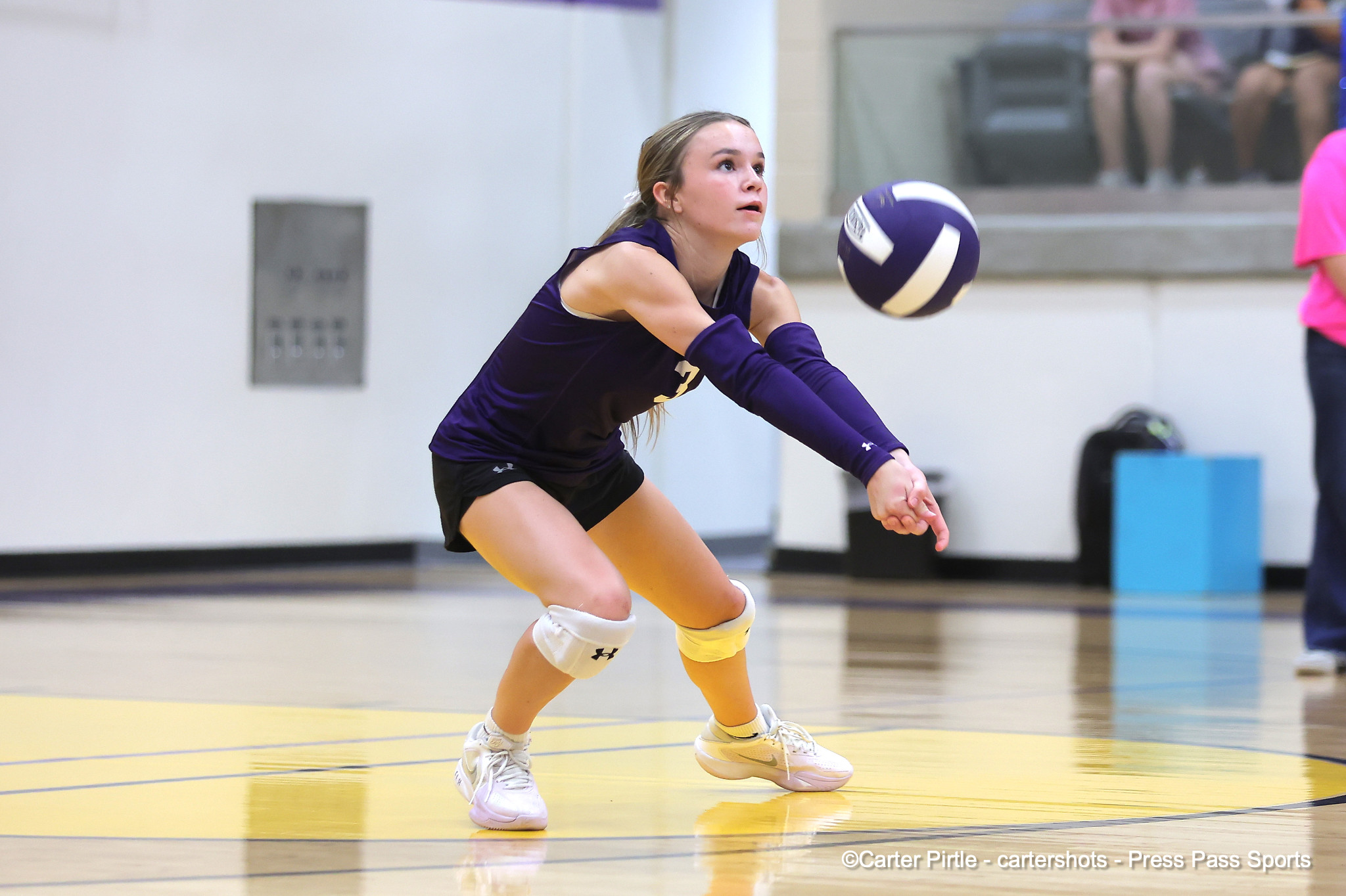 Female volleyball player in purple uniform dives to dig the ball on an indoor court, eyes on the ball, yellow court lines visible in foreground.
