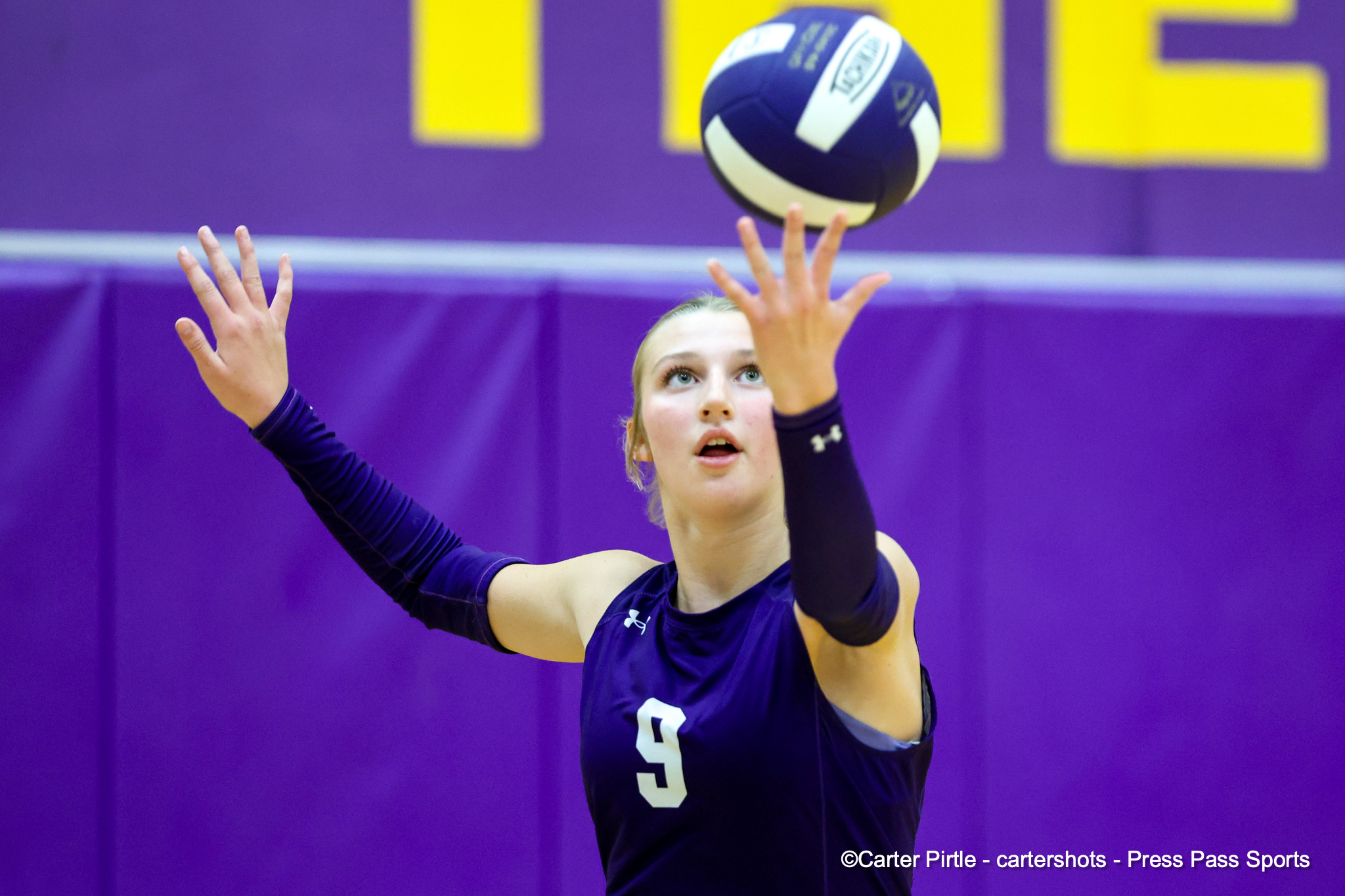 Female volleyball player in a purple uniform reaches up to hit a blue-and-white volleyball in a purple gym backdrop.
