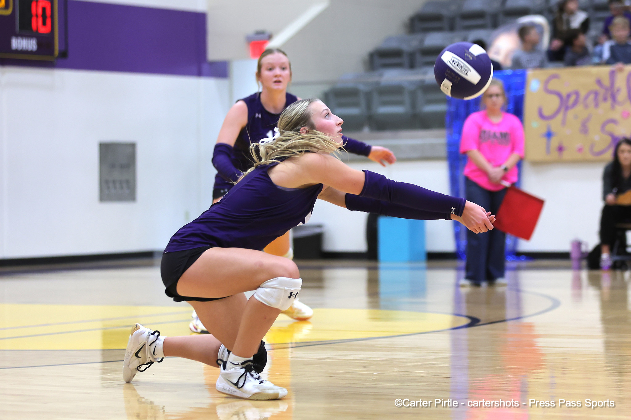 Two female volleyball players in purple dive for a ball on an indoor court; foreground player extends arms while the ball flies toward them, with spectators in the background.