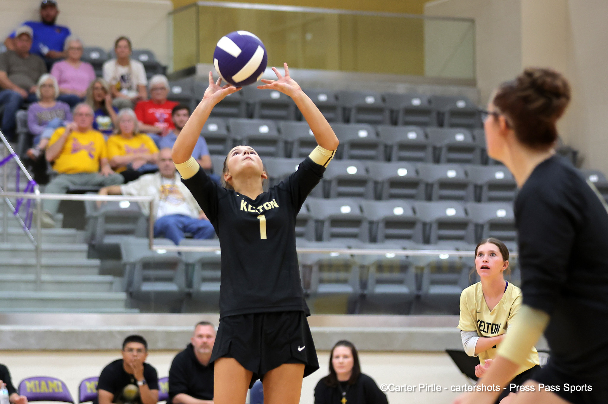 Volunteer? A female volleyball player in a black Kelton uniform (number 1) reaches up to touch a purple-and-white ball as she prepares to volley, with spectators seated in the gym bleachers behind her.