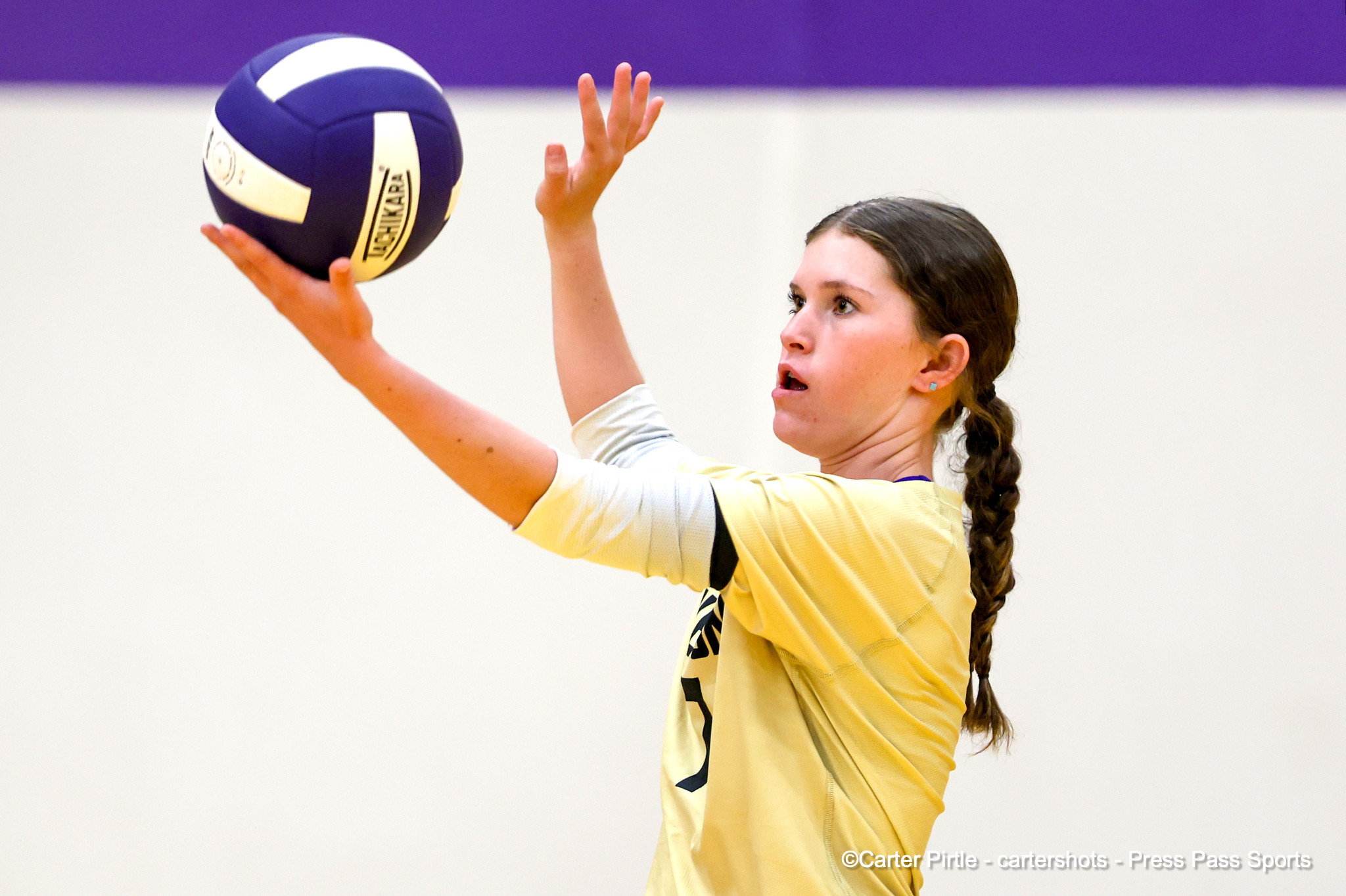 Female volleyball player with a braid and yellow jersey reaching to hit a blue volleyball.
