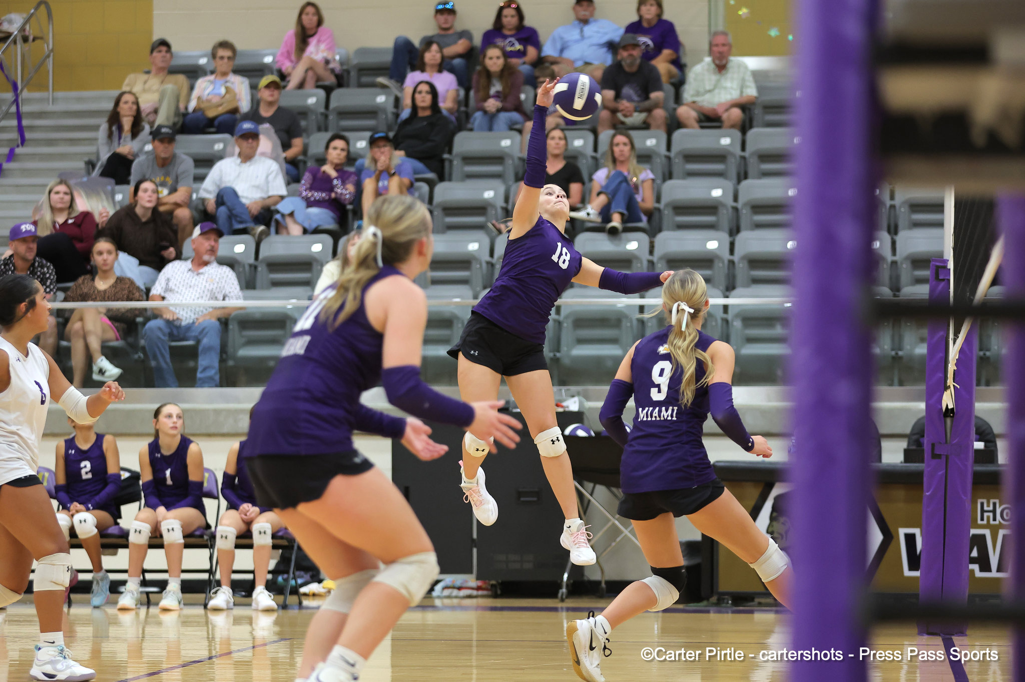 Female volleyball player in a purple jersey (#18) leaps to serve the ball during a match, with spectators watching from the stands behind her.