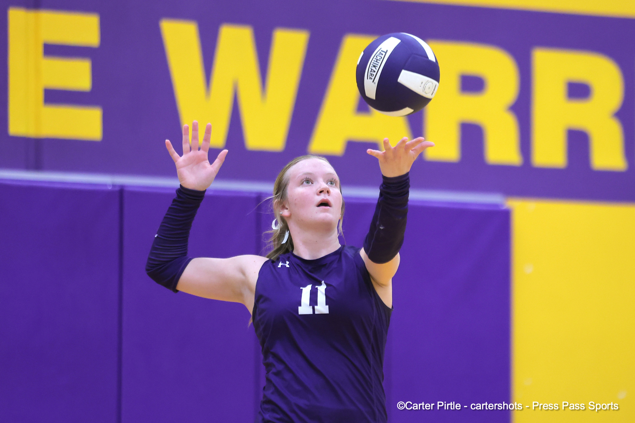 Female volleyball player in a purple jersey with number 11 reaches upward to contact a ball during a match against a purple-yellow court backdrop.
