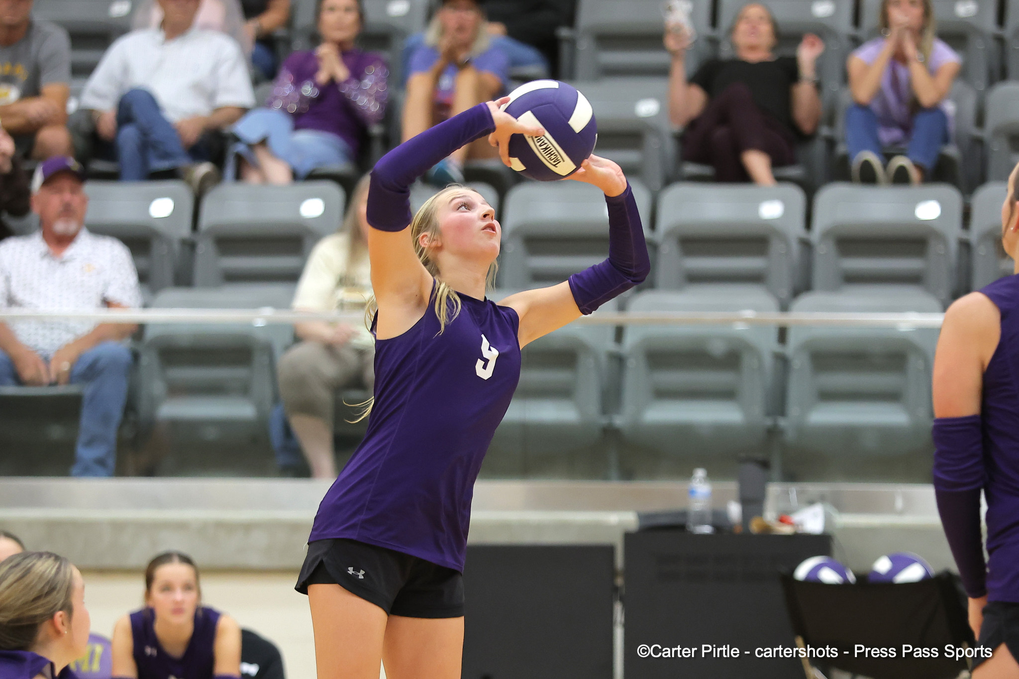 Female volleyball player in a purple uniform serving the ball in an indoor gym, with spectators in the stands behind her