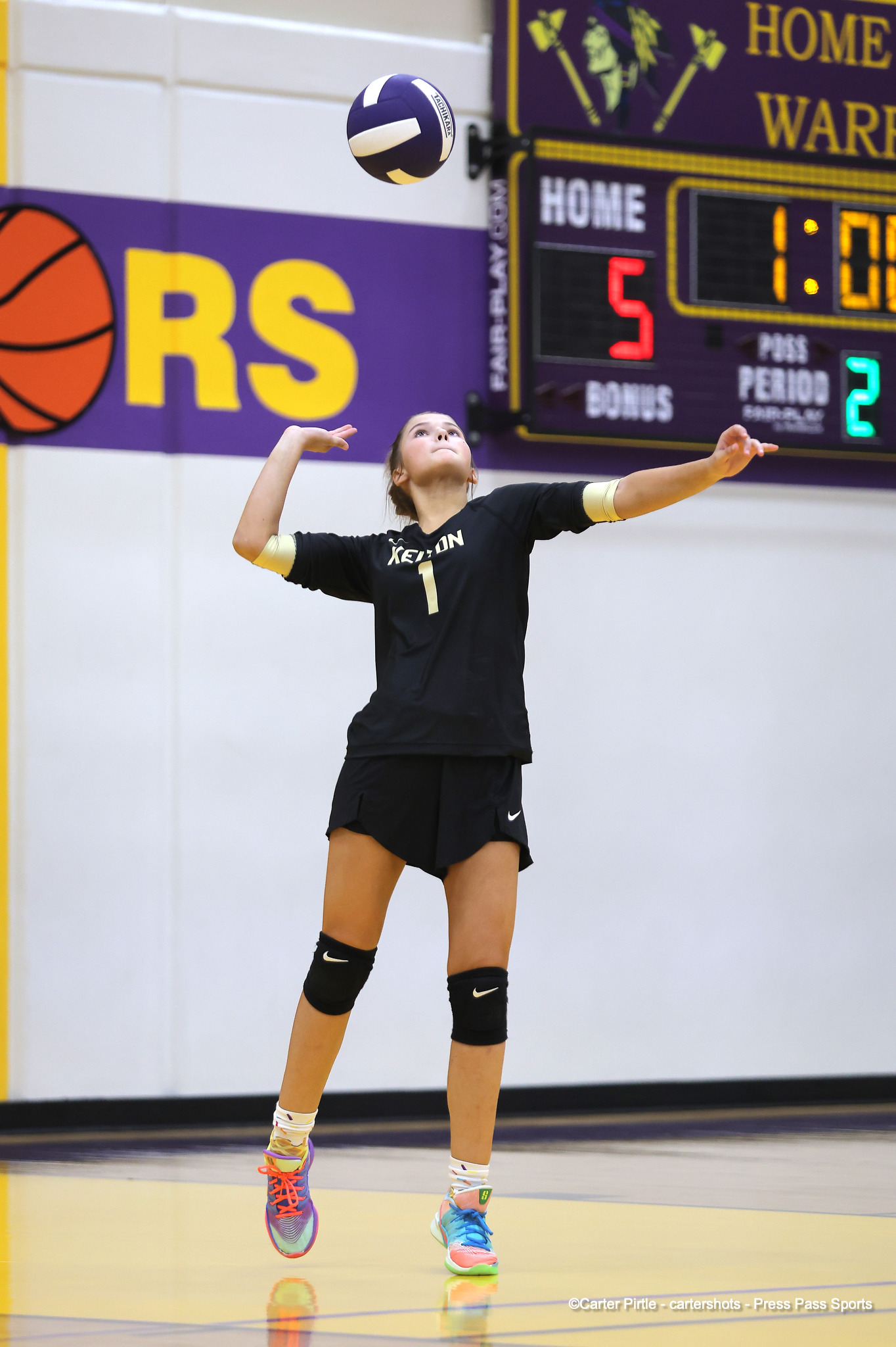 Female volleyball player in a black uniform with the number 1 serves the ball mid-air on an indoor court, with a scoreboard in the background.