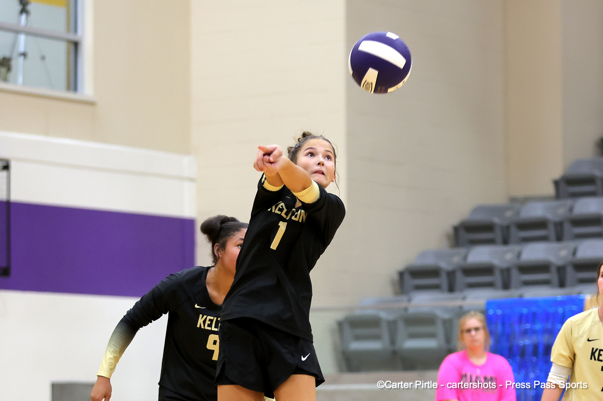 Female volleyball player in a black uniform reaching up to spike the ball in an indoor gym with purple walls and empty bleachers in the background.