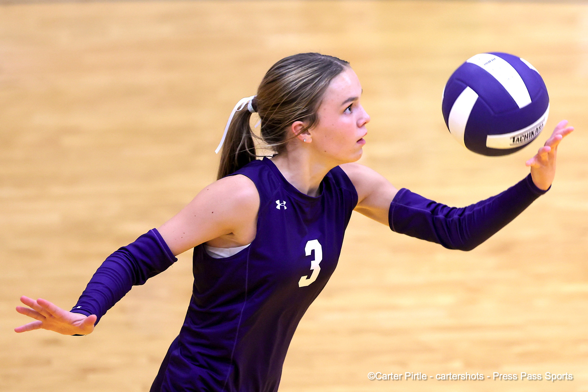 Female volleyball player in a purple uniform reaches for a purple-and-white ball during a tense rally on a gym court.