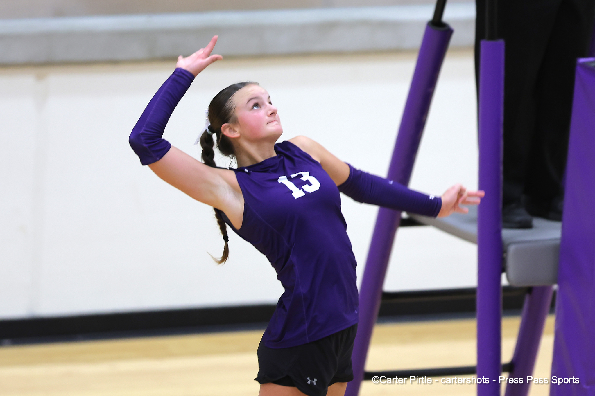 Female athlete in a purple uniform with number 13, arms raised mid-movement in a gym with purple equipment nearby