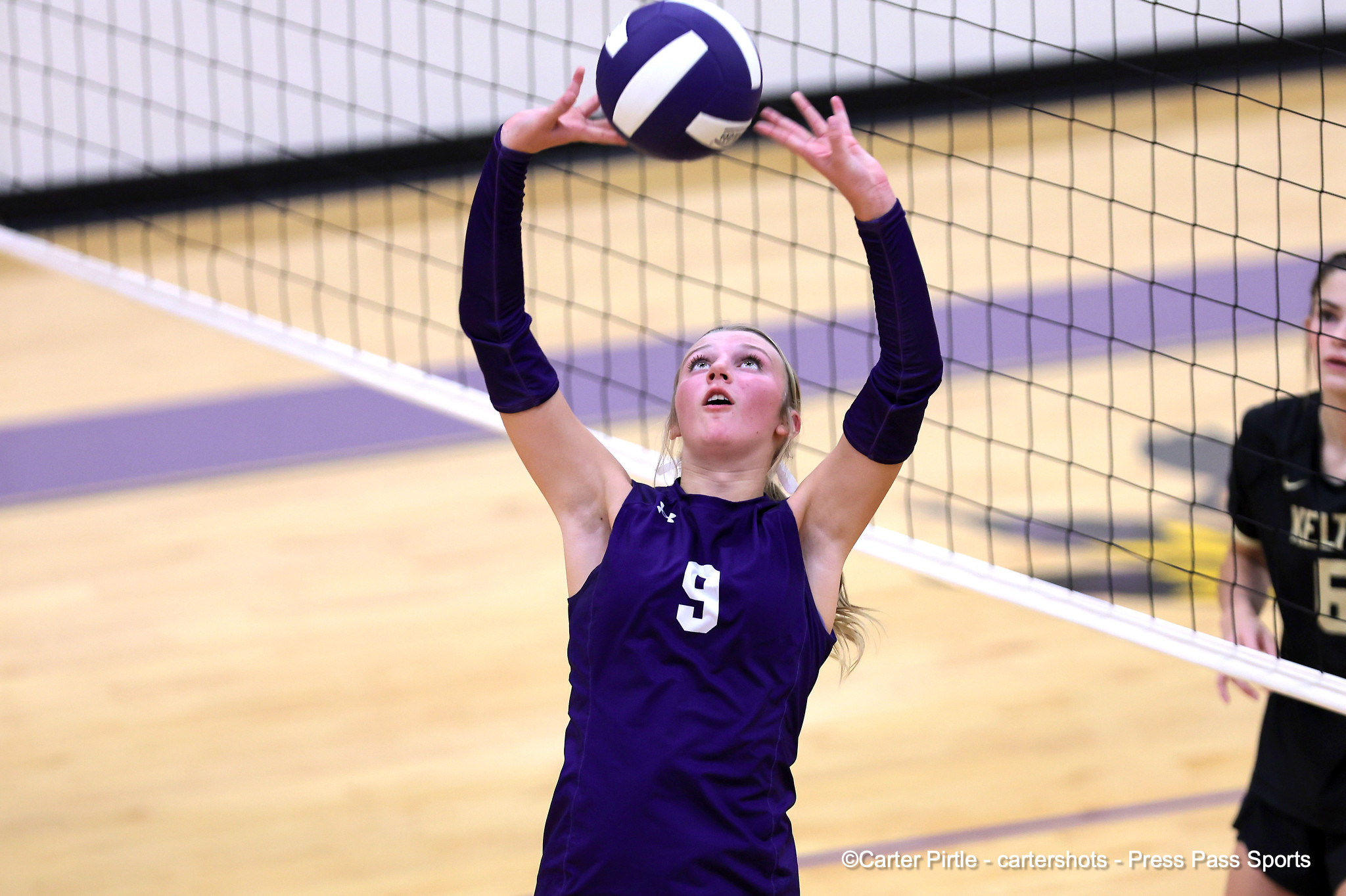 Female volleyball player in a purple jersey reaching up to set the ball over the net during a match
