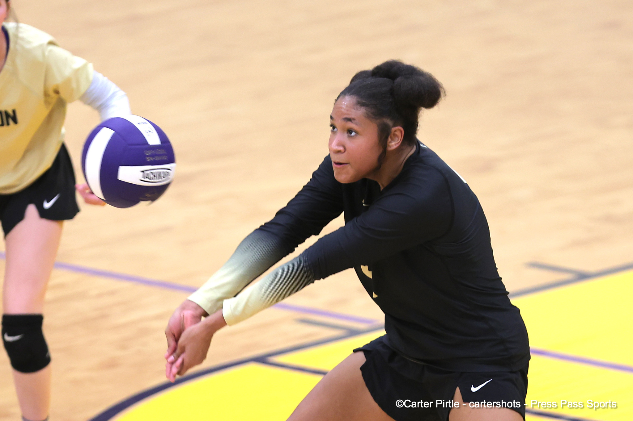 Female volleyball player in black lunges toward a purple-and-white ball during a game in a gymnasium.
