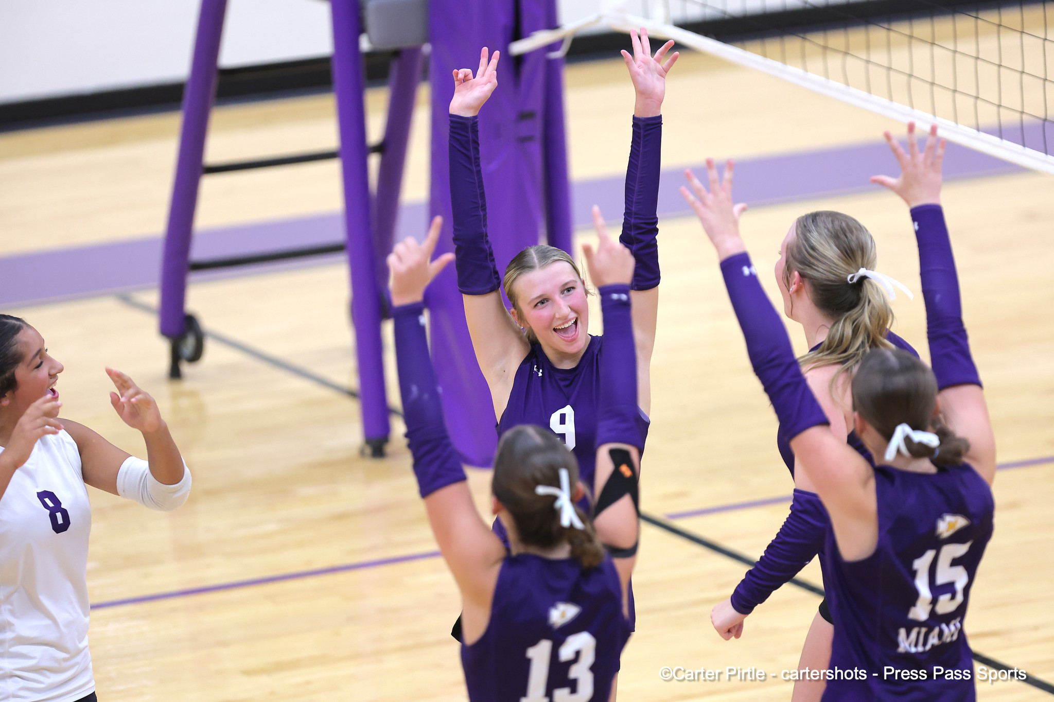 Girls' volleyball team in purple jerseys celebrate with raised hands on a gym court