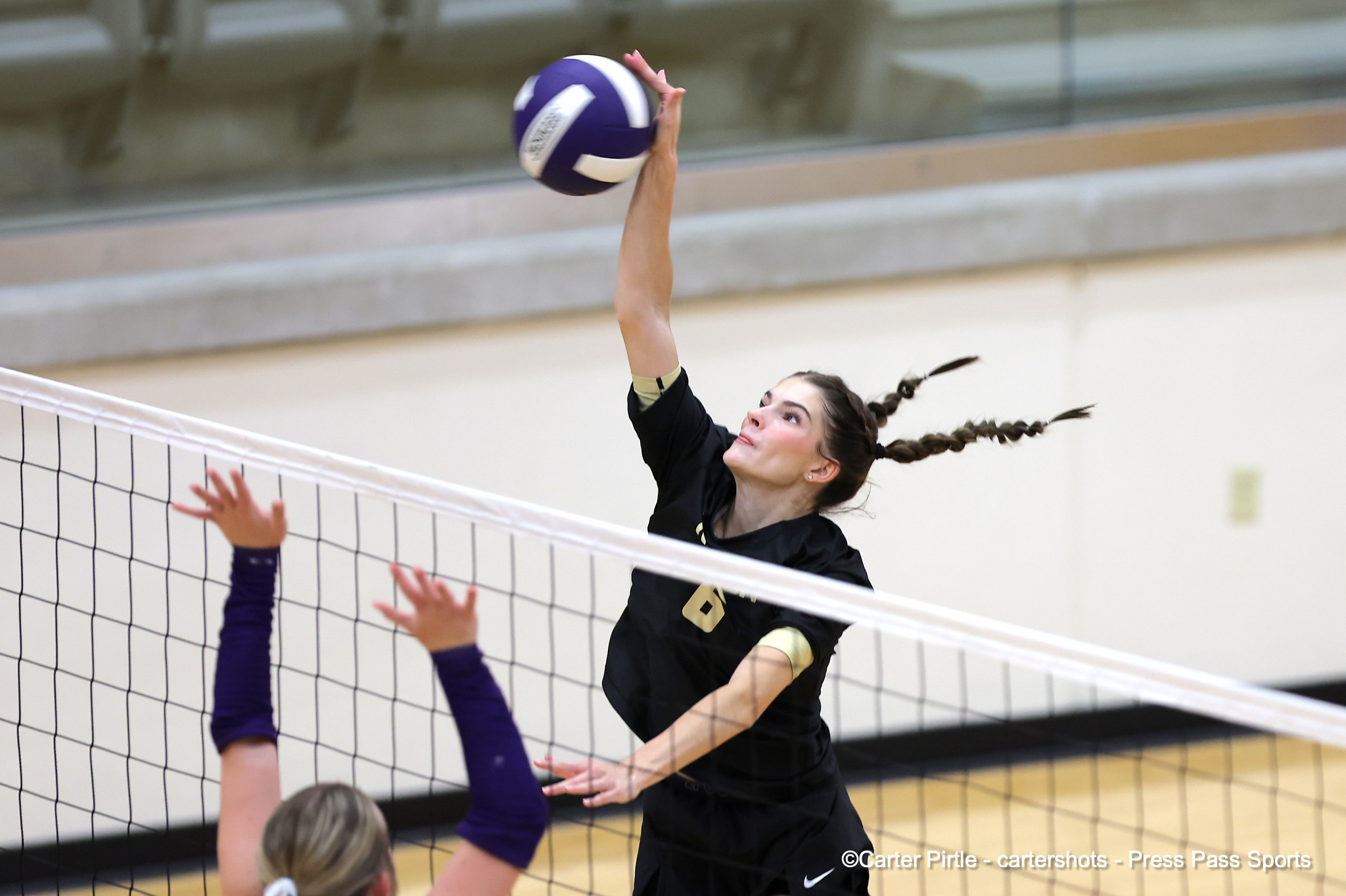 Female volleyball player jumping to spike the ball over the net in an indoor gym, dark uniform, braided hair.