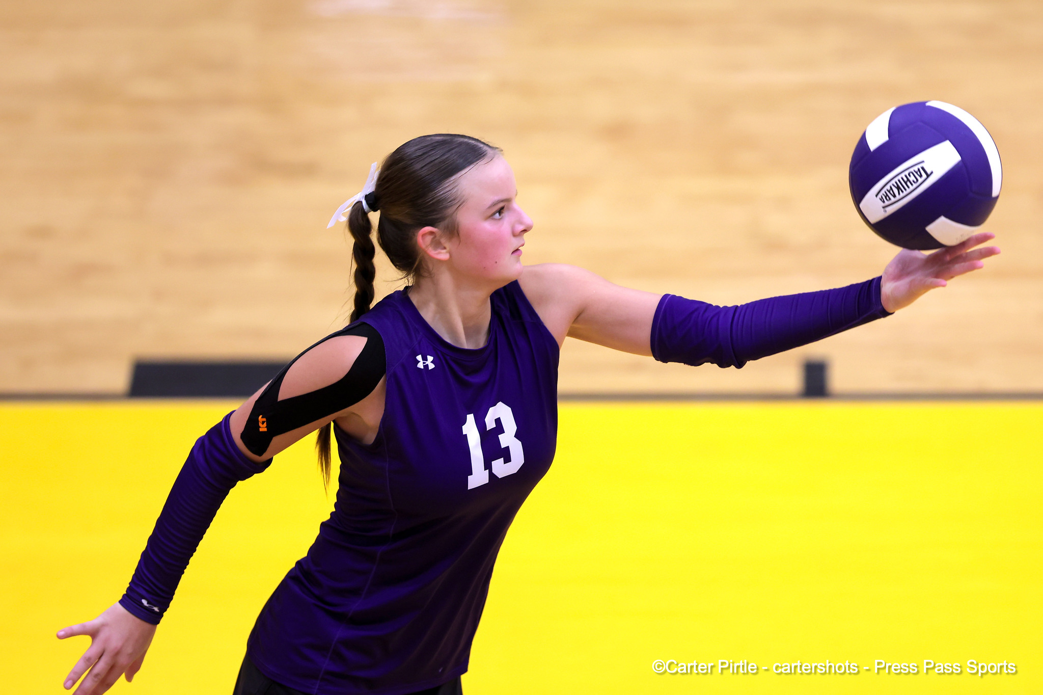 Female volleyball player in purple jersey #13 extends arm to hit a ball in a gym with a yellow wall.