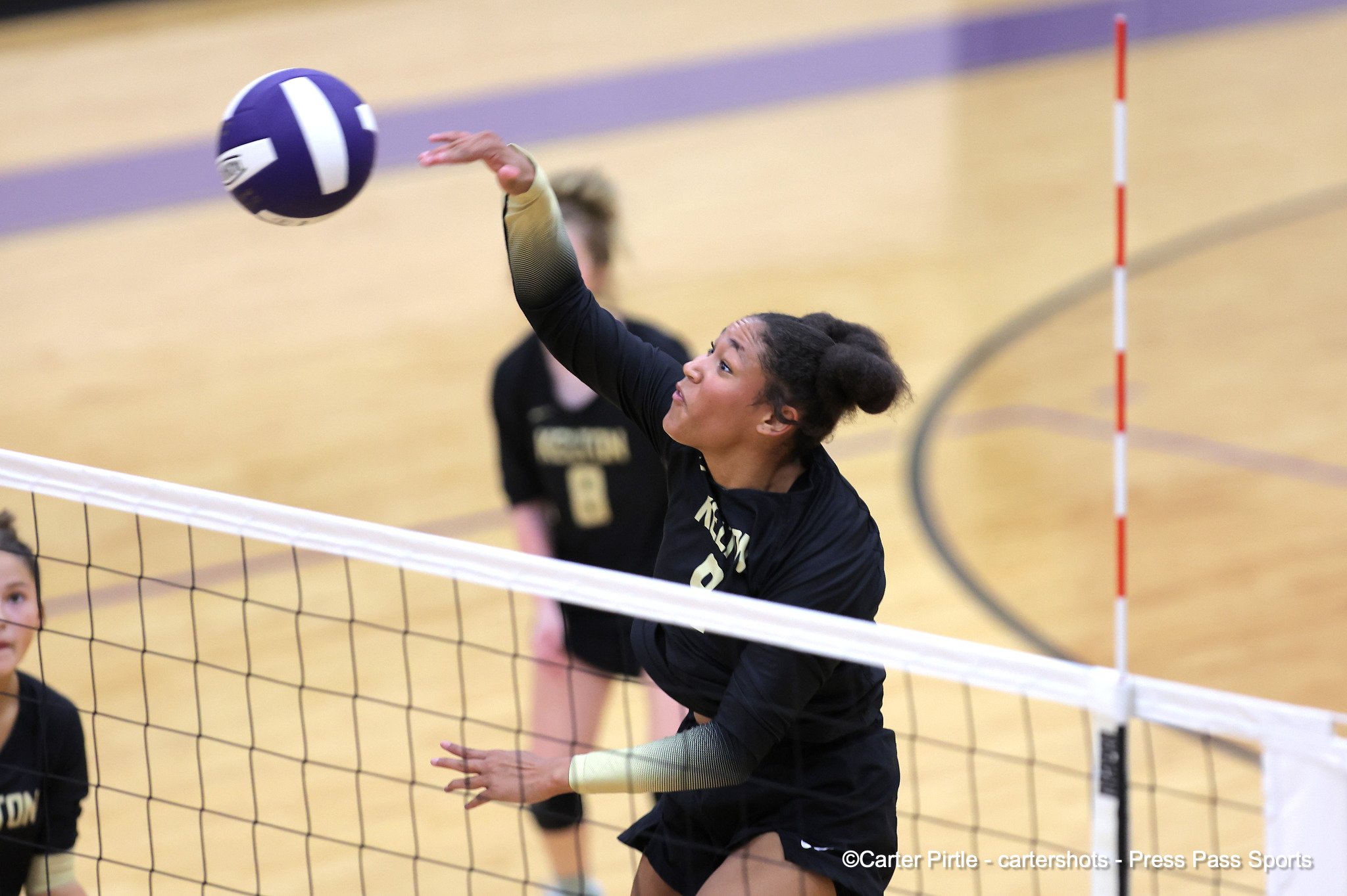 Female volleyball player in a black uniform spikes a purple volleyball over the net during a match in a gymnasium.
