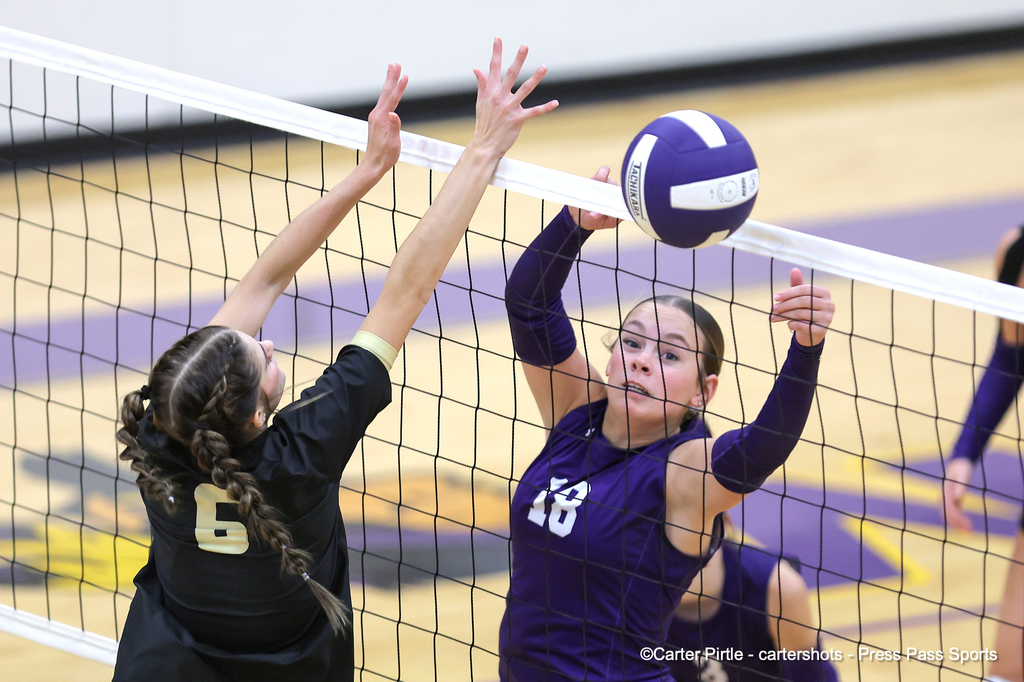 Two female volleyball players reach over the net to contact a purple-and-white ball during a match on a gym court.