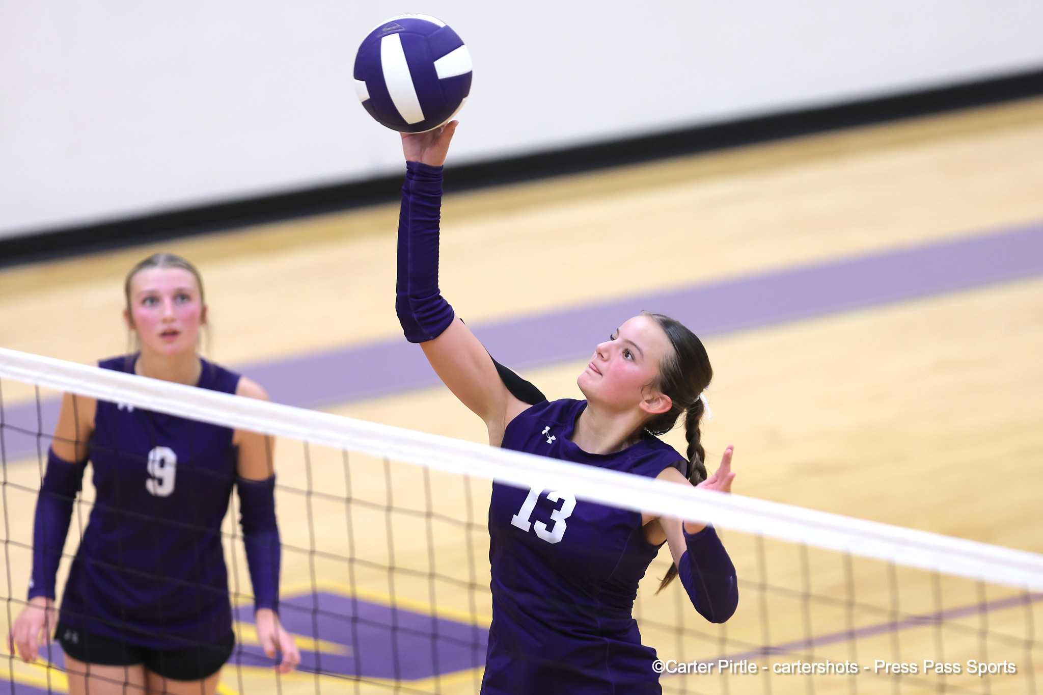 Female volleyball players in purple uniforms at the net; one player reaches up to spike the ball.