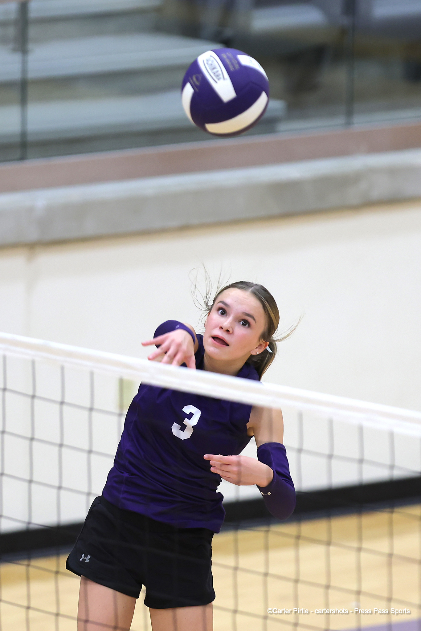 Female volleyball player in a purple jersey with number 3 reaching toward the ball at the net in a gym.