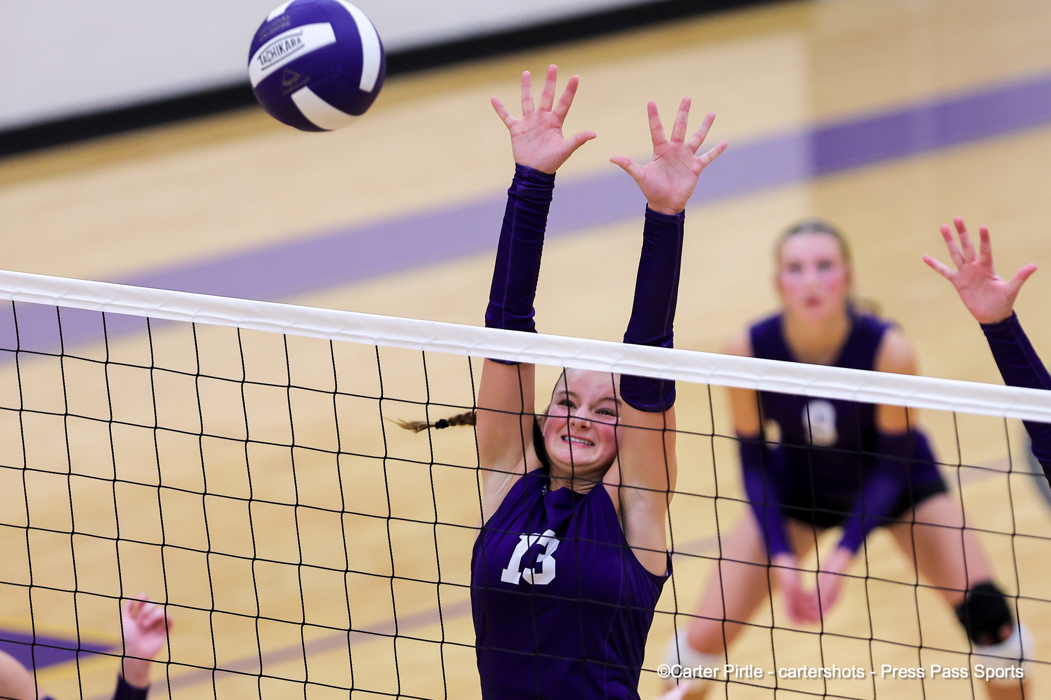 Volleyball players at the net jumping to block a ball, wearing purple uniforms.