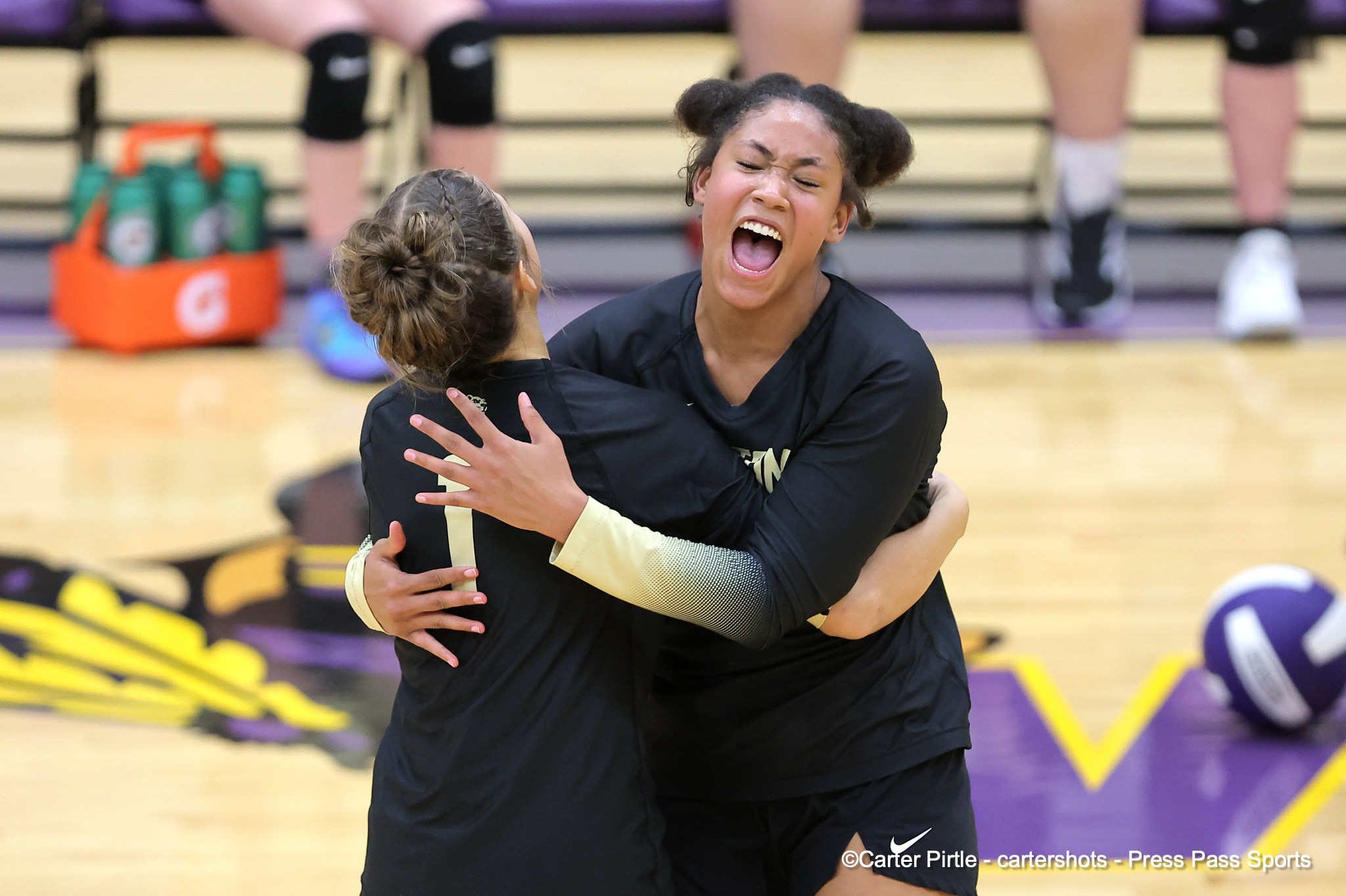 Two teammates hug in a gym, one with mouth open in celebration and a volleyball nearby on the court.