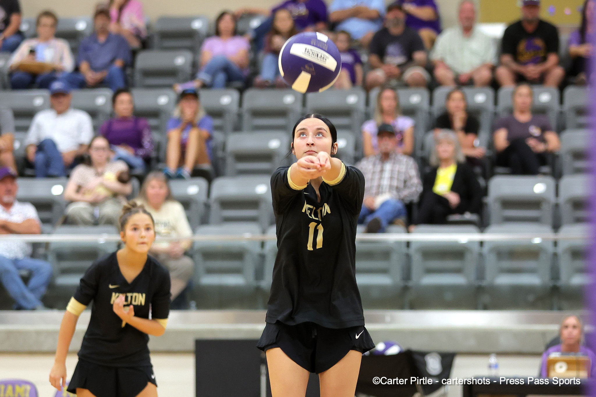 Female volleyball player in black uniform bumps or passes the ball while teammates watch in an indoor arena with spectators behind her.