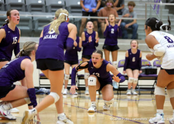 20260922-Kelton-Miami-VB-043 | Press Pass Sports Women’s volleyball game in a gym; players in purple uniforms cheer as one dives for the ball near the floor, with a white-uniform opponent on the right.