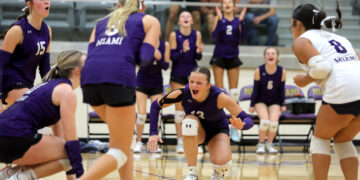 Women’s volleyball game in a gym; players in purple uniforms cheer as one dives for the ball near the floor, with a white-uniform opponent on the right.