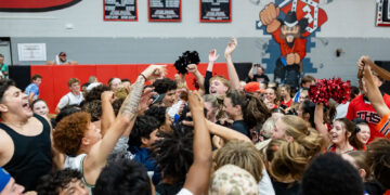 Crowd of students cheering and raising hands in a gym during a pep rally, with red wall padding and banners in the background.
