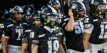 Raiders football players in black and white uniforms gather in a tunnel, foreground player wearing number 19 and neon green mouthguard.