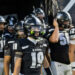 CHS-RHS-FB-2 1 | Press Pass Sports Raiders football players in black and white uniforms gather in a tunnel, foreground player wearing number 19 and neon green mouthguard.
