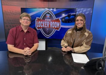 IMG_7697 | Press Pass Sports Two TV hosts sit at a desk in a studio, with a large screen showing the 'The Locker Room' logo behind them.