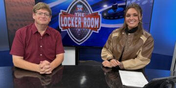 Two TV hosts sit at a desk in a studio, with a large screen showing the 'The Locker Room' logo behind them.