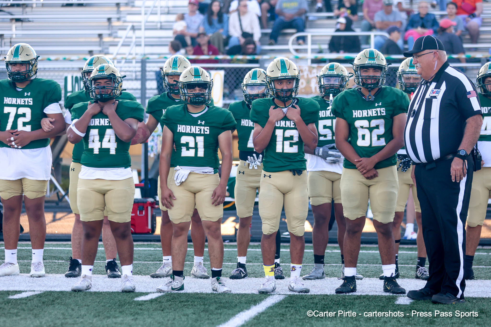 Group of Hornets football players in green jerseys and tan shorts lined up on the field with a referee on the right during a pre-game moment.