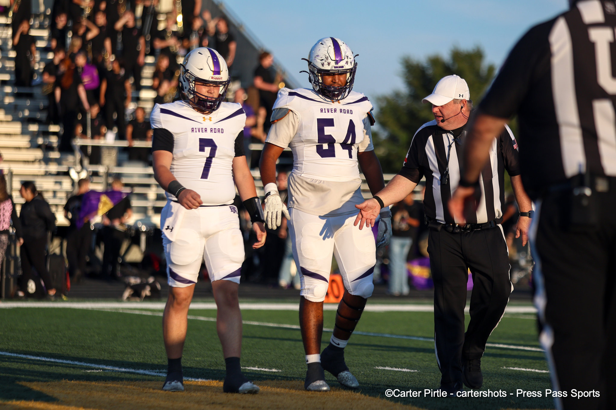Two River Road football players walk toward the sideline on a sunny field, #7 on the left and #54 in the center, with a referee nearby and a blurred crowd in the stands behind them.