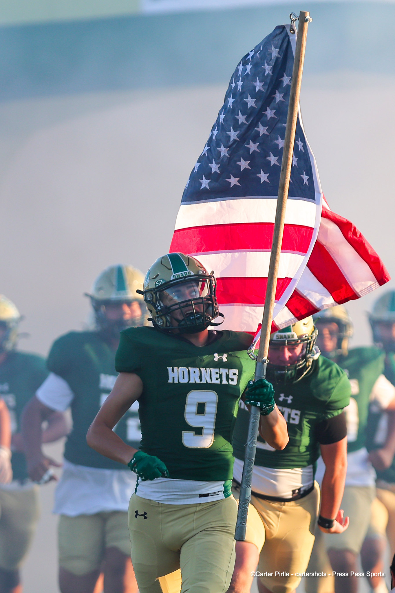 Hornets football players run forward as a large American flag waves overhead.