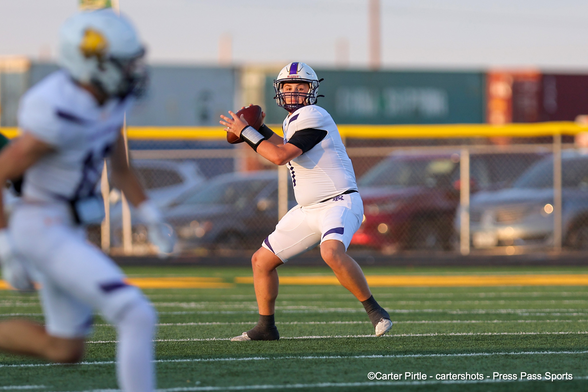 Quarterback in white uniform holds a football, preparing to throw on a sunny football field.