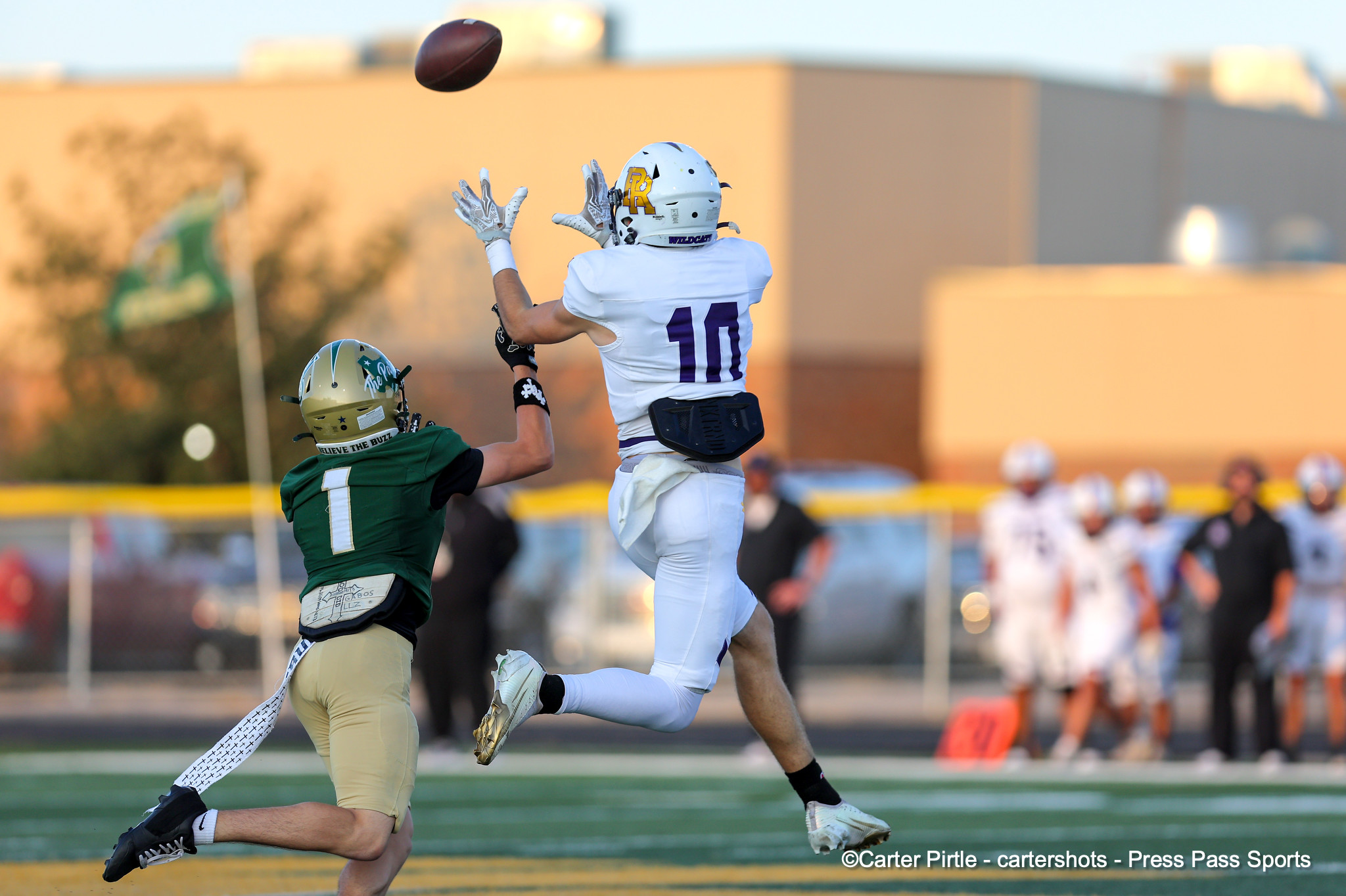 Two football players leap for a pass as the ball flies overhead on a sunny field, with a blurred crowd in the background.