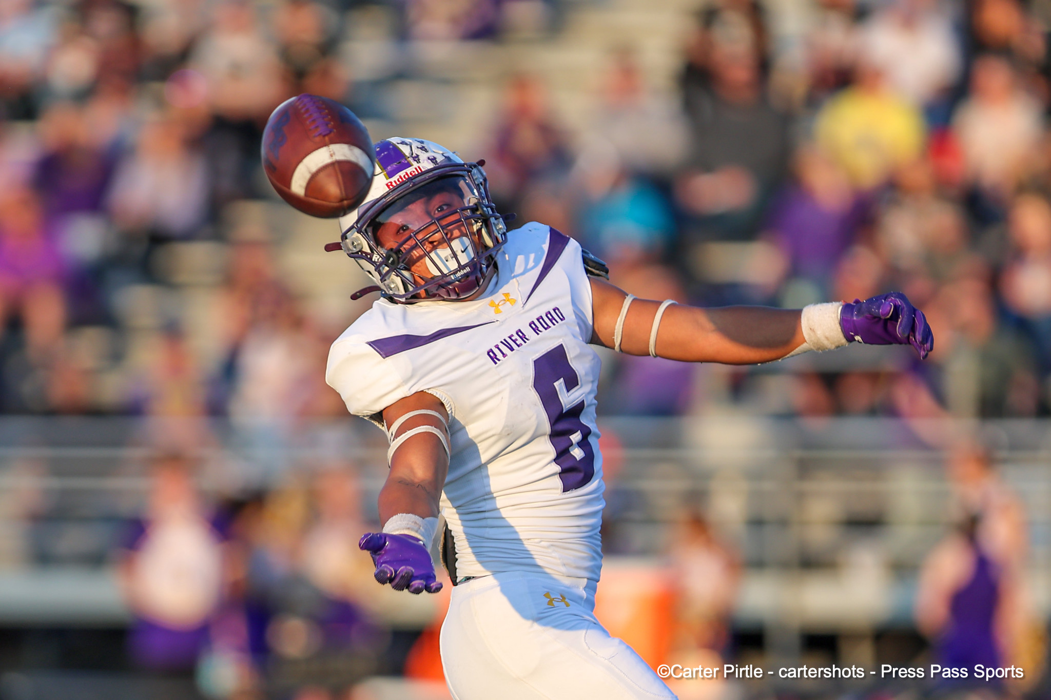 Football player in white and purple uniform reaching for a midair football with a blurred crowd in the stands behind.