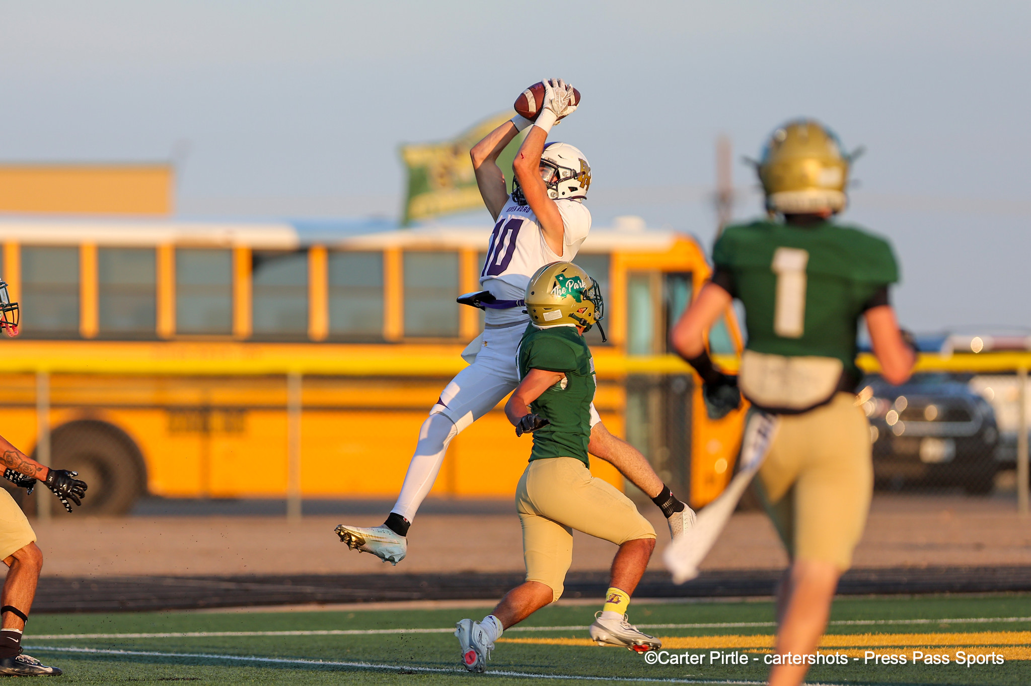 Football player in white jumps to catch a pass while a defender in green reaches up nearby on a sunny sideline field. A yellow school bus is blurred in the background.