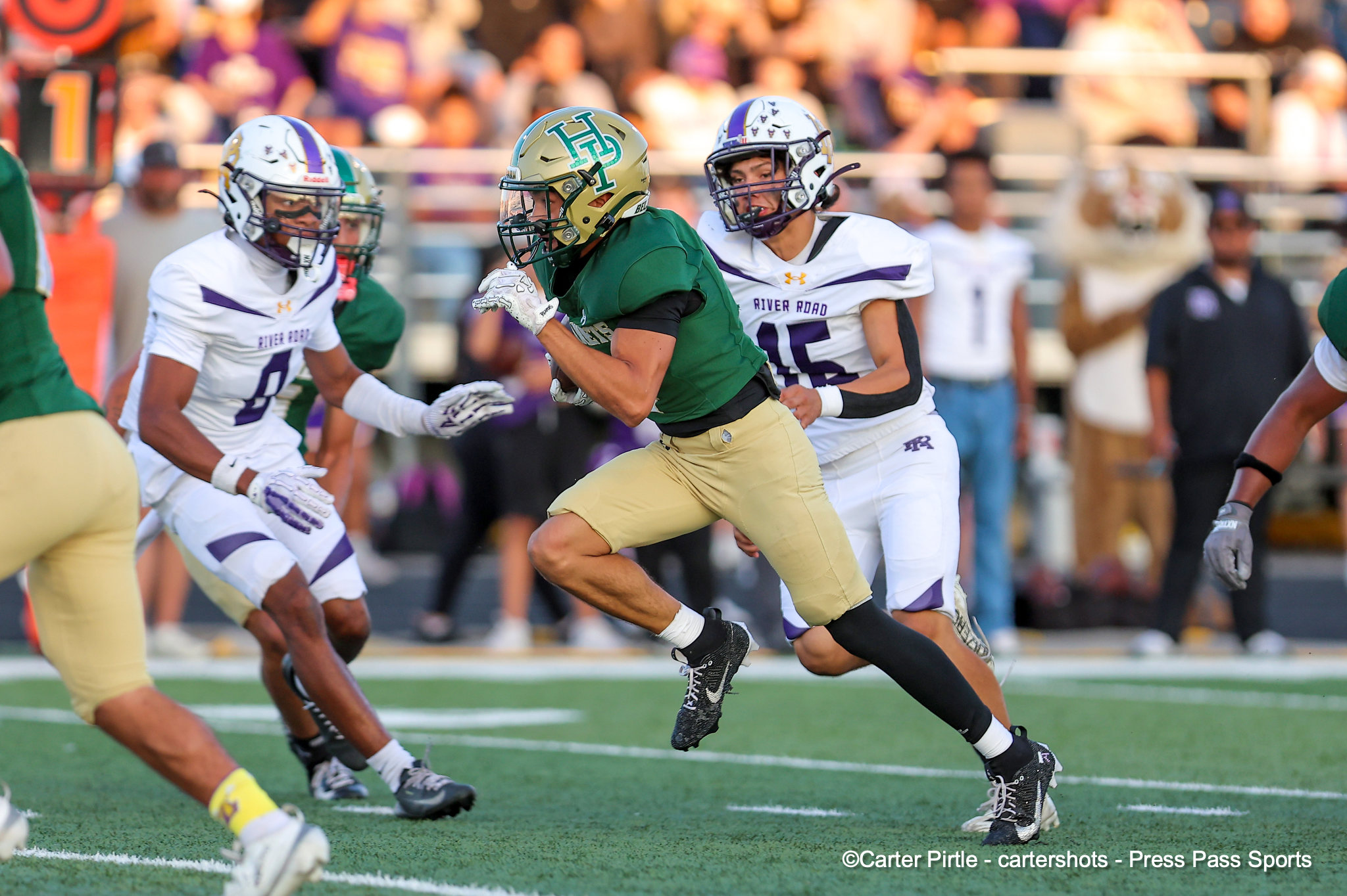 A football player in green jersey runs with the ball while teammates and defenders chase on a grassy field during a game.