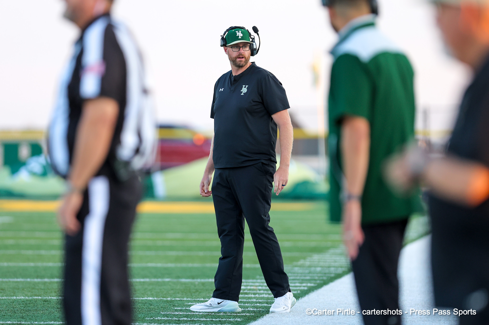 Coach wearing headset and black athletic wear stands on a football field, looking to the side near the sideline.