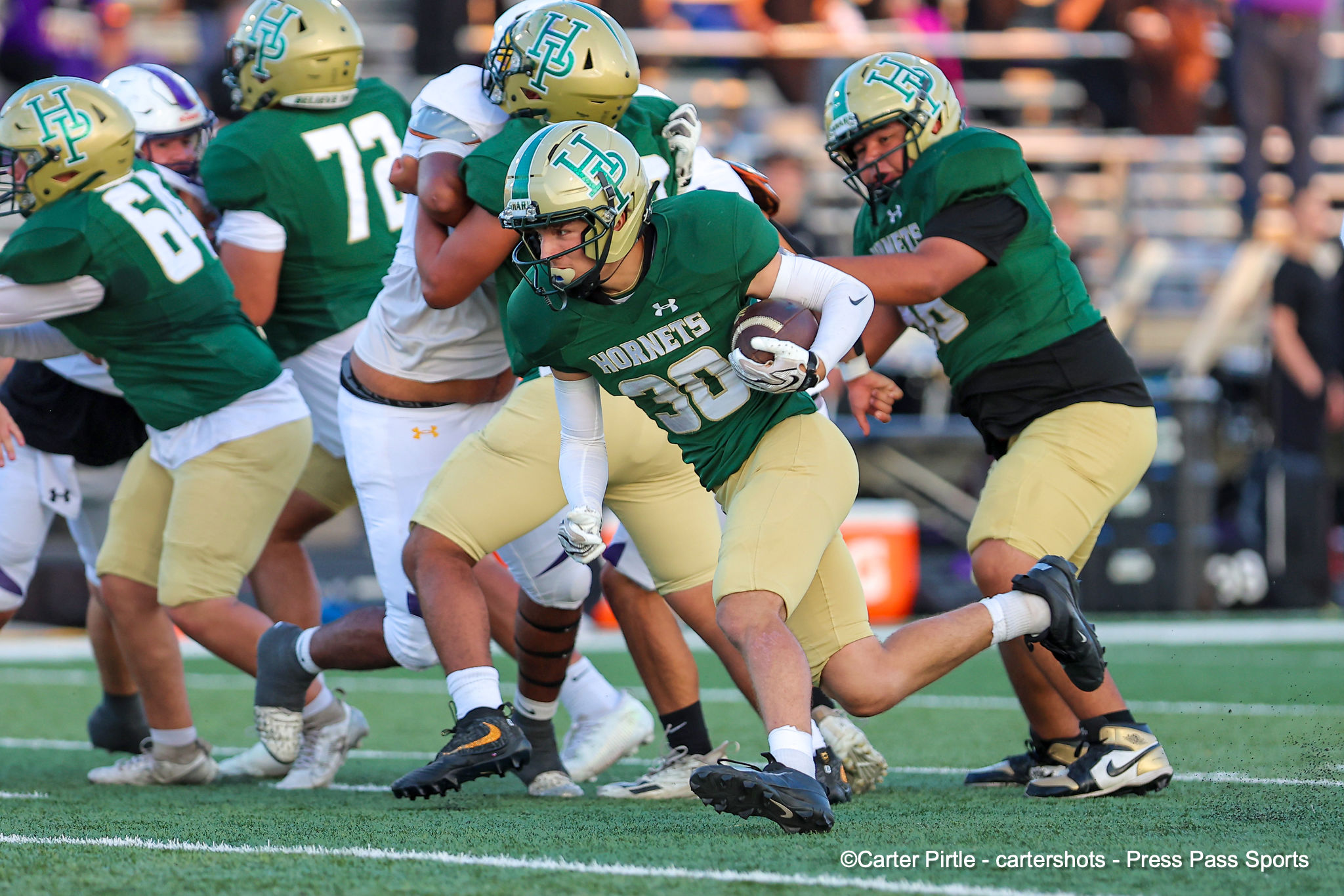 Green-and-gold Hornets players sprint with the ball during a football game on a turf field, front-and-center ball carrier in action.